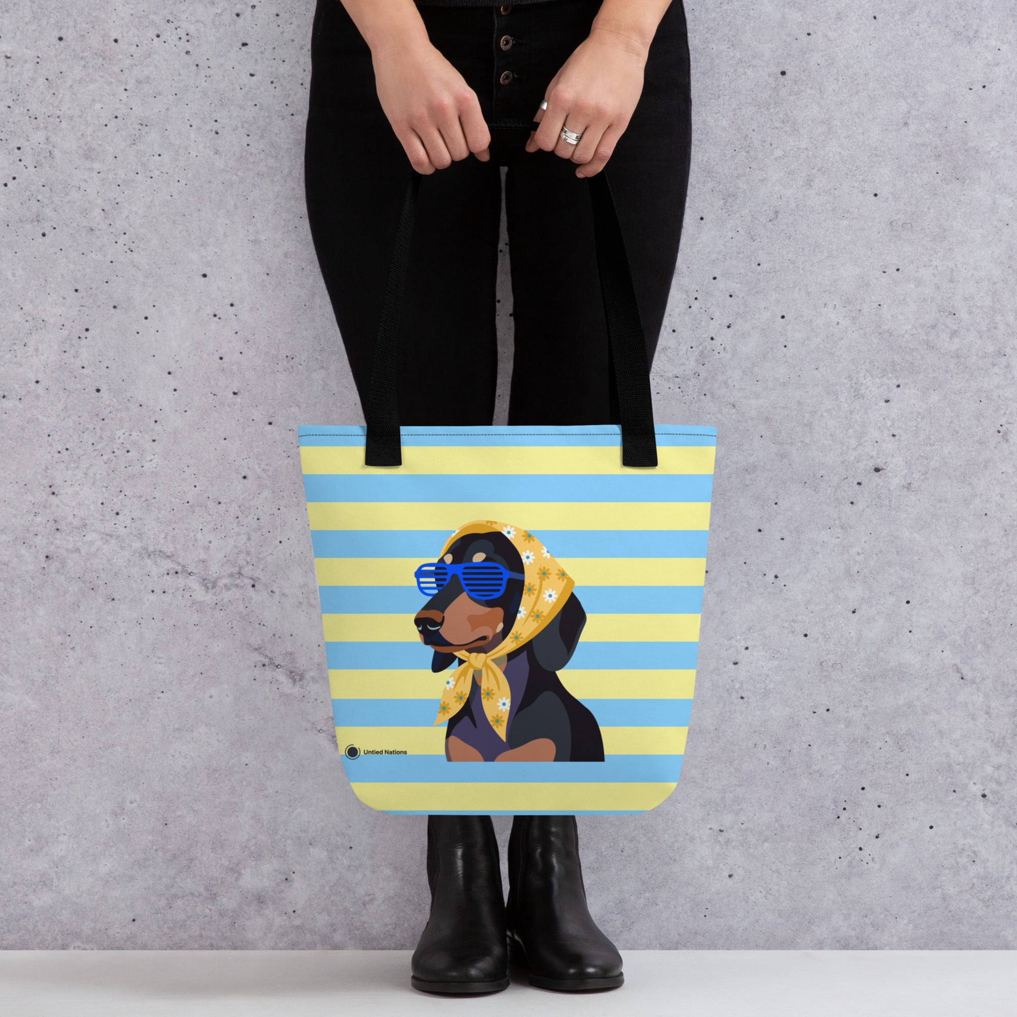 A person wearing black pants and black boots holds a tote bag with a cartoon dachshund wearing blue sunglasses and a yellow bandana, set against a gray concrete wall.