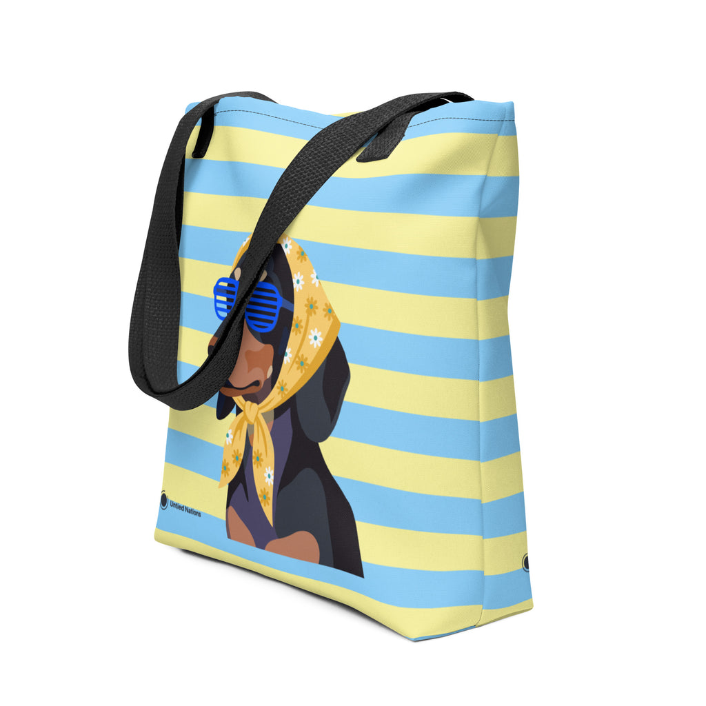 A canvas tote bag with a light blue and pale yellow horizontal stripe pattern, featuring a cartoon illustration of a black dachshund wearing a yellow bandana and blue sunglasses. The bag has black webbed handles and is shown on a plain white background.