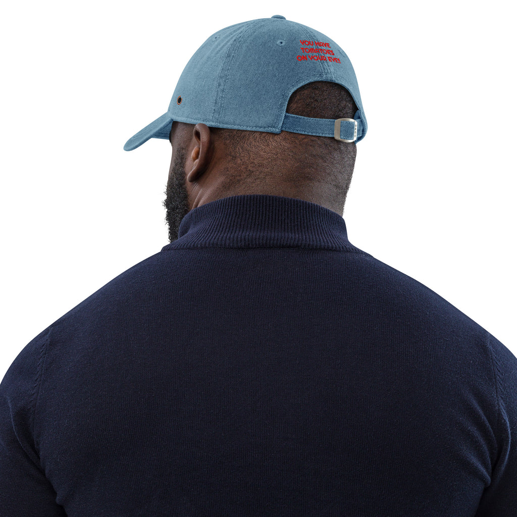 A man viewed from behind wearing a light blue denim baseball cap with red embroidered text and a navy blue ribbed turtleneck sweater, isolated on a white background.