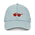 A light blue denim baseball cap featuring an embroidered pair of tomatosunglasses on the front, set against a plain white background.