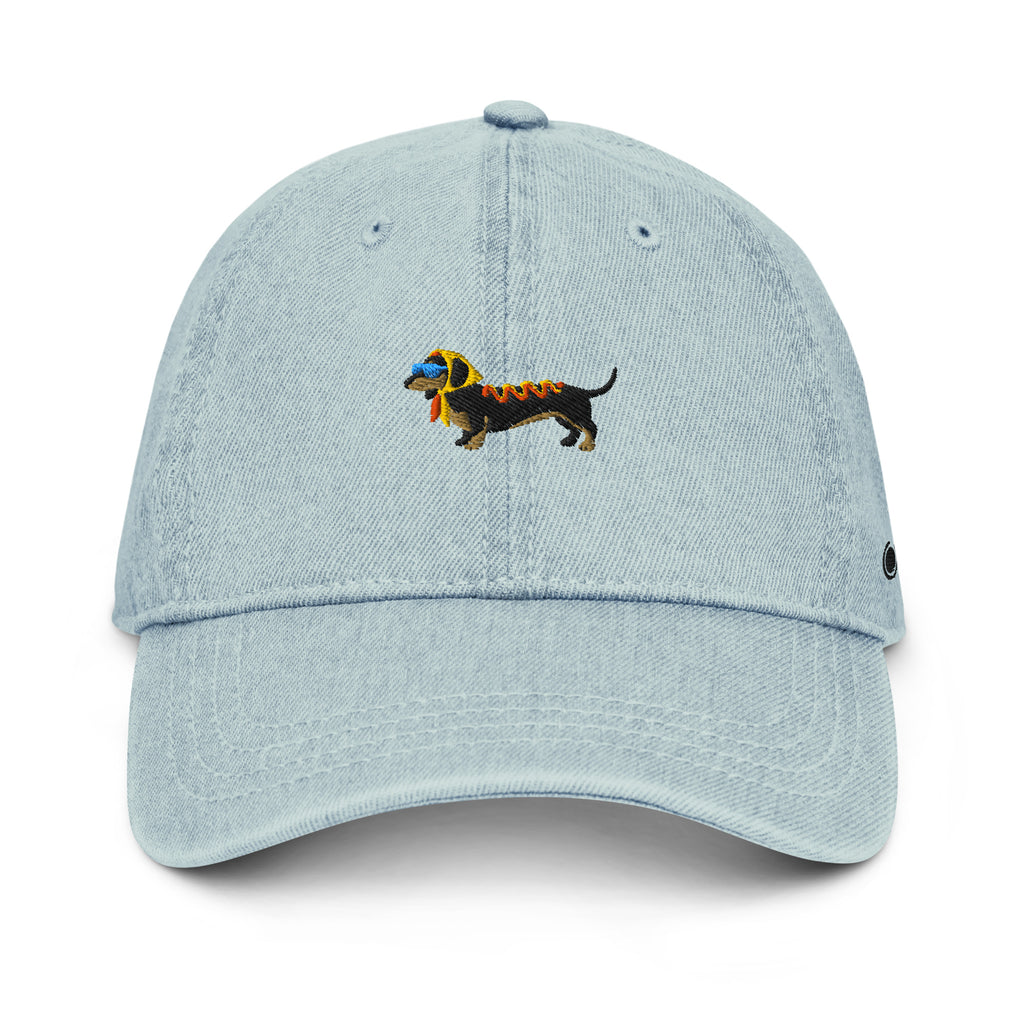 A light blue denim baseball cap featuring an embroidered dachshund dog on the front. The dog is black with a yellow headscarf, a pair of light blue sunglasses, and ketchup and mustard on its back.