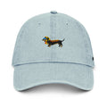 A light blue denim baseball cap featuring an embroidered dachshund dog on the front. The dog is black with a yellow headscarf, a pair of light blue sunglasses, and ketchup and mustard on its back.