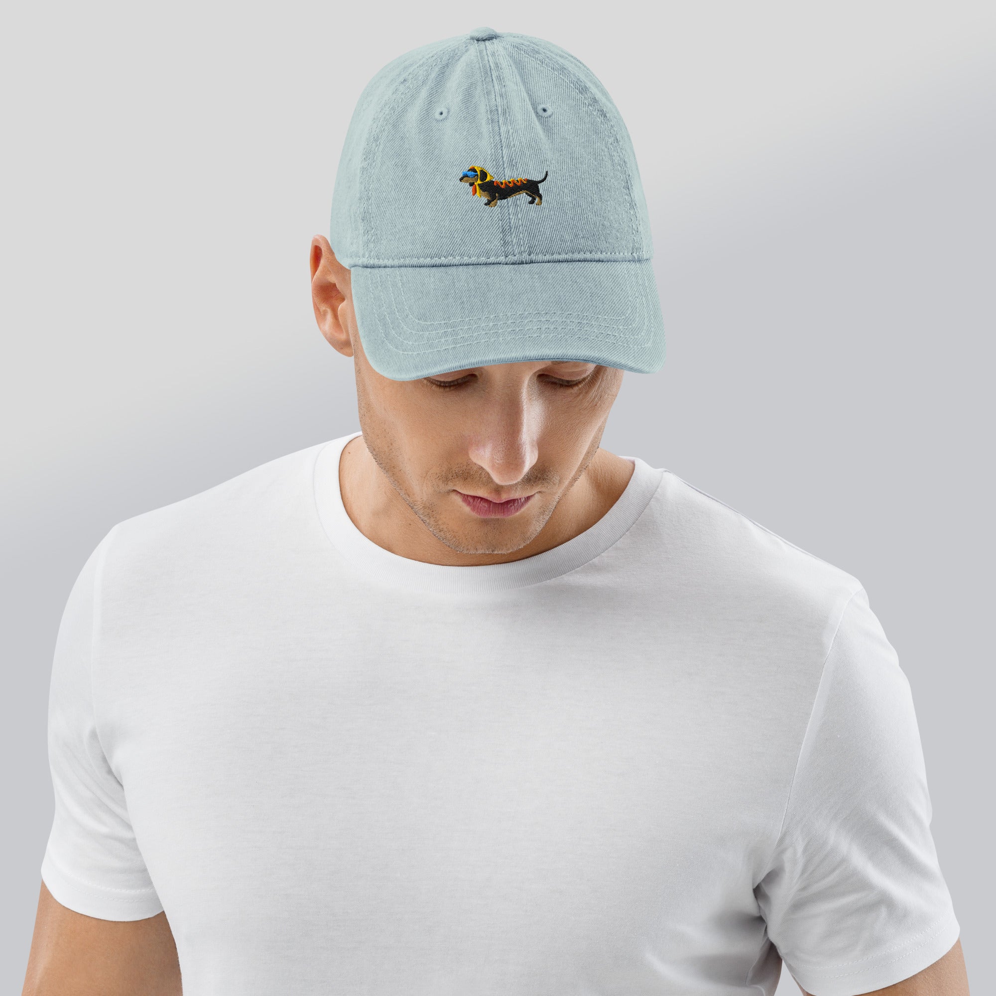 Man wearing a light blue denim baseball cap featuring an embroidered dachshund dog on the front. The dog is black with a yellow headscarf, a pair of light blue sunglasses, and ketchup and mustard on its back.