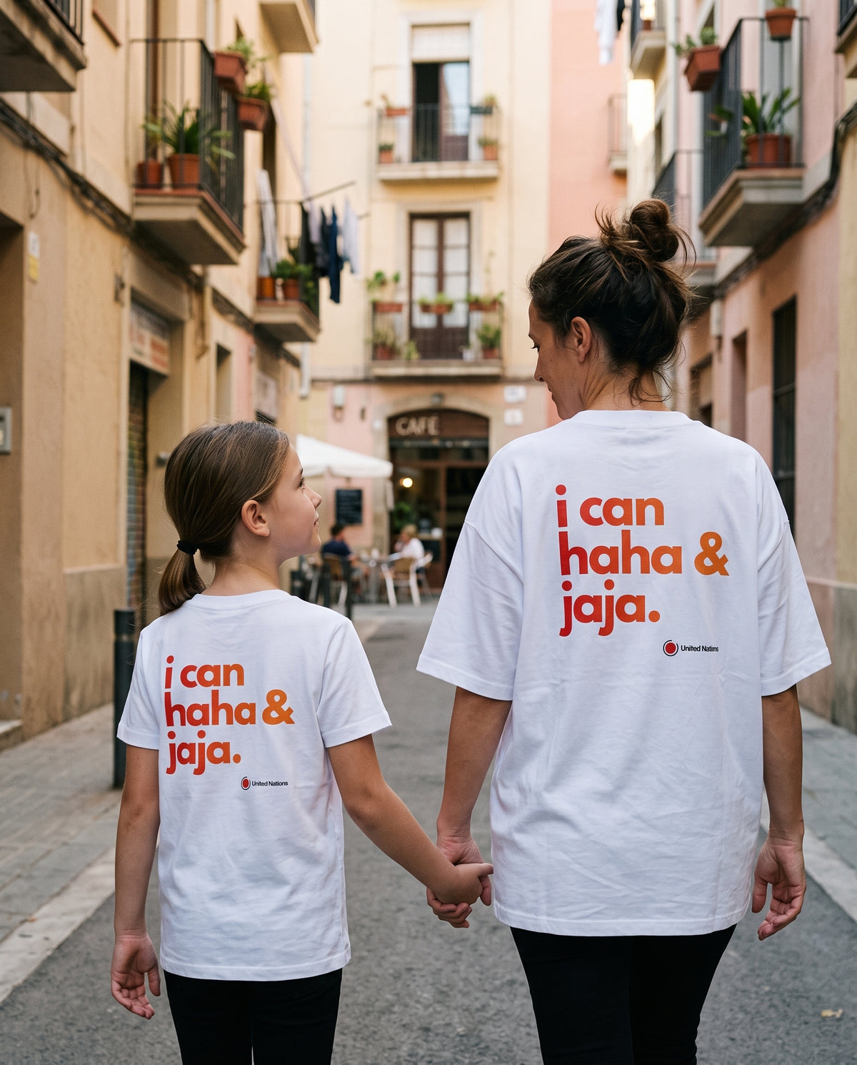 A woman and a girl walking hand-in-hand down a narrow European street, both wearing matching white graphic T-shirts with orange text that reads 'i can haha & jaja.' on the back.