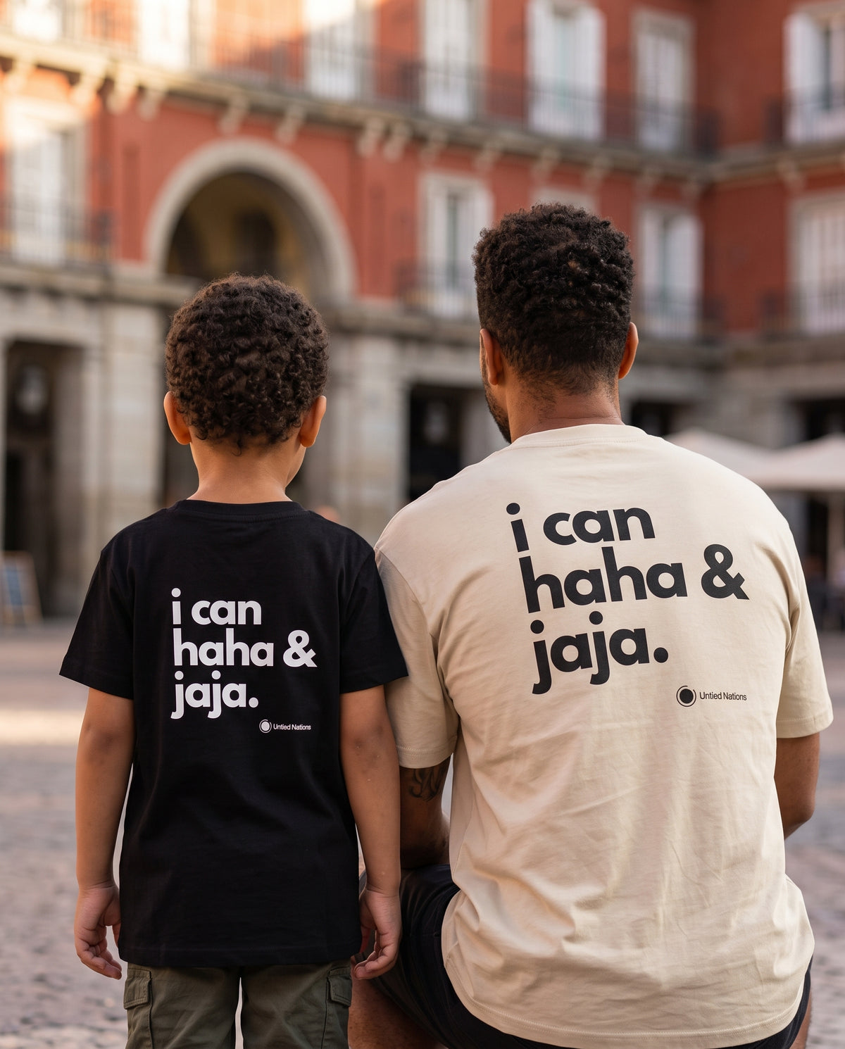 A man and a child seen from behind wearing matching graphic t-shirts with the text 'i can haha & jaja.' printed on the back, set against a blurred outdoor plaza background.
