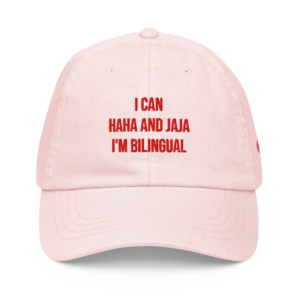 A light pink dad hat baseball cap with red embroidered text on the front that reads 'I CAN HAHA AND JAJA I'M BILINGUAL'.