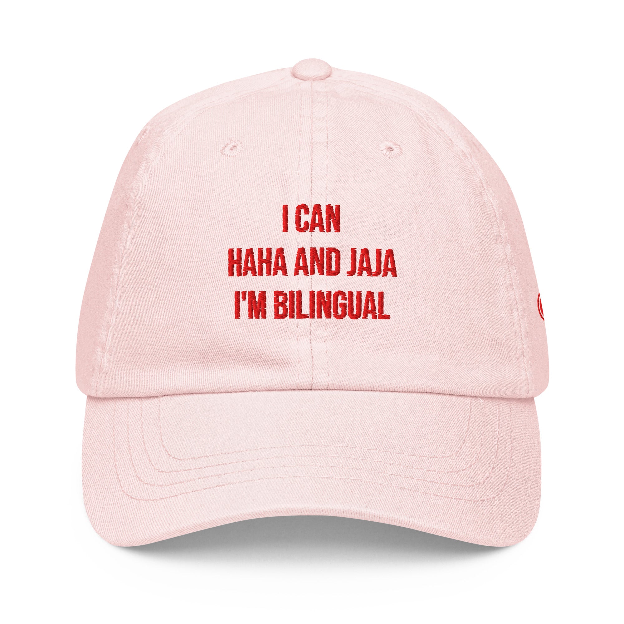A light pink dad hat baseball cap with red embroidered text on the front that reads 'I CAN HAHA AND JAJA I'M BILINGUAL'.