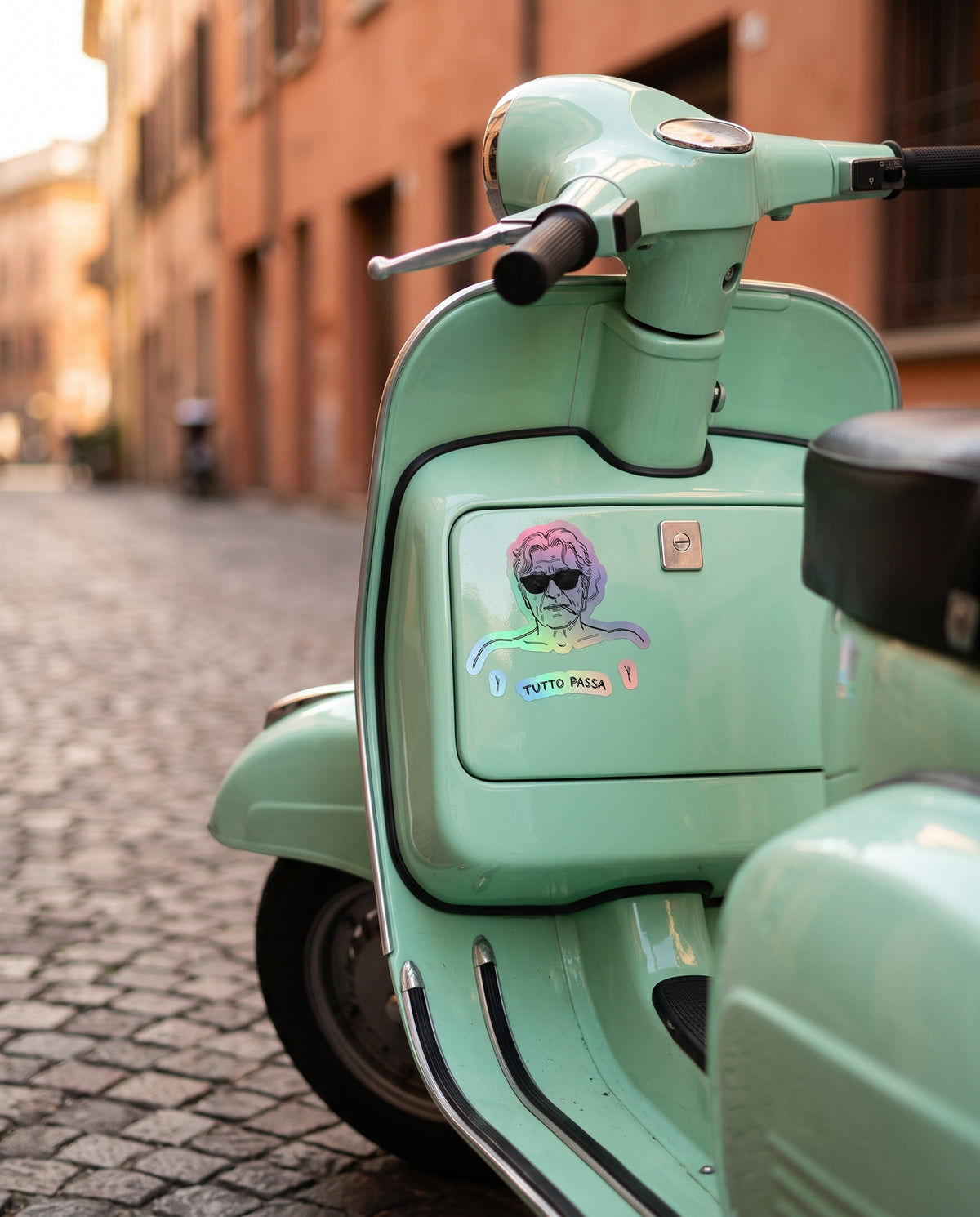 A mint green Vespa scooter parked on a cobblestone street in front of warm-toned buildings. A colorful sticker with a stylized face and the text 'tutto passo' is applied to the side panel.