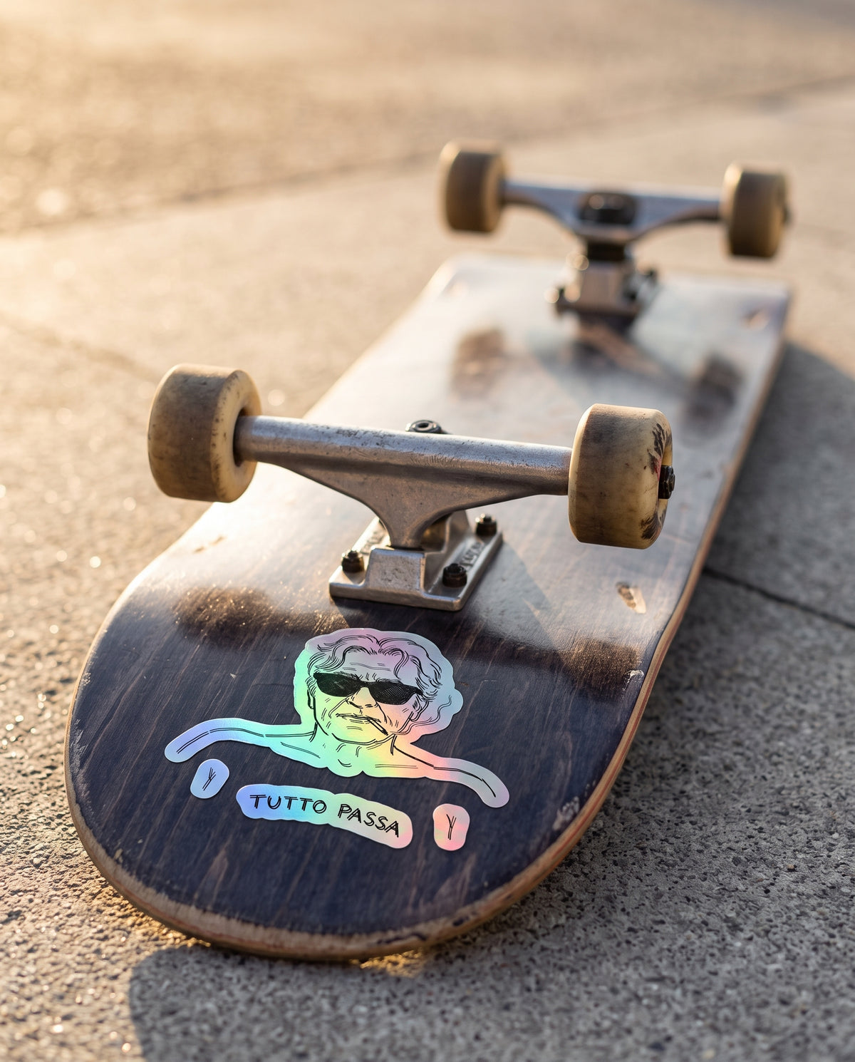 A close-up of a skateboard deck lying on concrete pavement, showing a dark worn wood surface with a colorful holographic sticker of a man in sunglasses and the text 'TUTTO PASSA'. The skateboard has metal trucks and tan wheels, photographed in warm natural sunlight.
