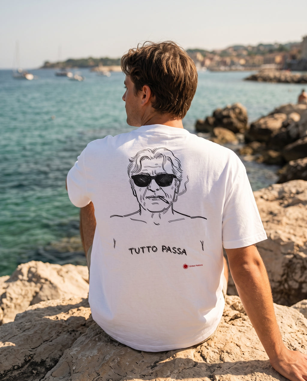 A man sitting on rocks by the sea, viewed from behind, wearing a white t-shirt with a black line art graphic of a man wearing sunglasses and the text 'TUTTO PASSA' printed below.