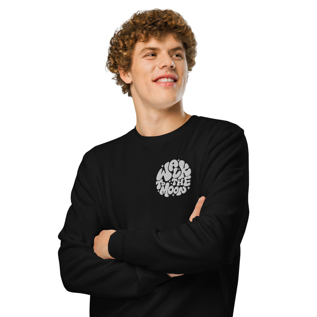 A smiling young man with curly brown hair wearing a black long-sleeve crewneck sweatshirt with a white circular graphic on the left chest that reads 'WALK TO THE MOON'. He is standing with his arms crossed against a solid white background.