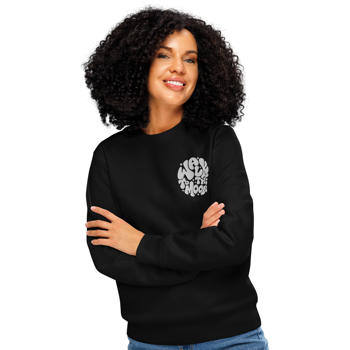A smiling woman with curly black hair wearing a black crewneck sweatshirt with a white circular graphic on the left chest that reads 'Walk to the moon'. She is posed with her arms crossed, set against a solid white background.