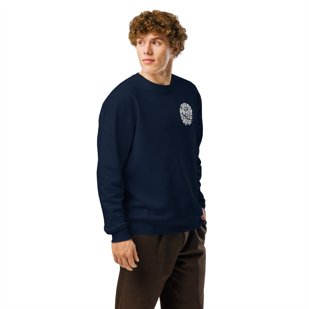 A man with curly brown hair wearing a navy blue crewneck sweatshirt with a small white graphic on the left chest, paired with dark brown pants, against a solid white background.