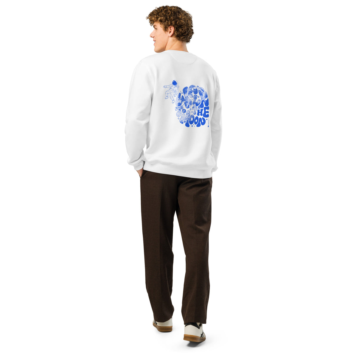 A man shown from the back wearing a white crewneck sweatshirt with a blue graphic print on the back, paired with dark brown pants and white sneakers, against a plain white background.