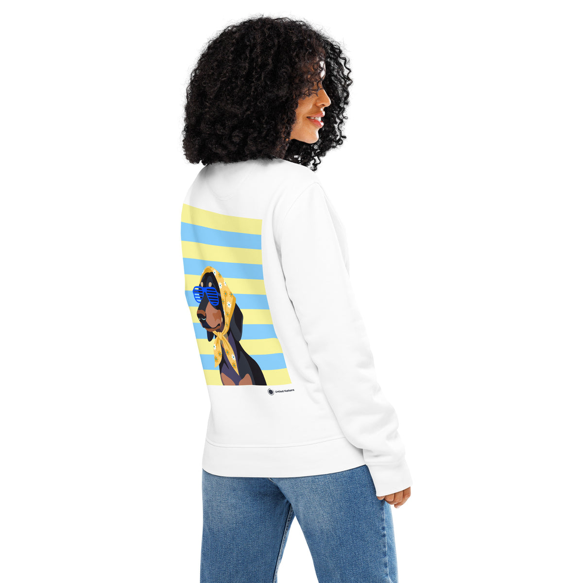 A woman shown from the back wearing a white sweatshirt with a large graphic print on the back, featuring a woman in a yellow headscarf and blue sunglasses against a yellow and blue striped background. She is also wearing blue jeans, set against a plain white studio background.