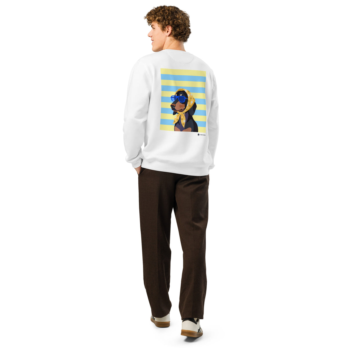 A man shown from the back wearing a white crewneck sweatshirt with a large graphic print of a cartoon dog on a yellow and blue striped background, paired with dark brown trousers and white sneakers.