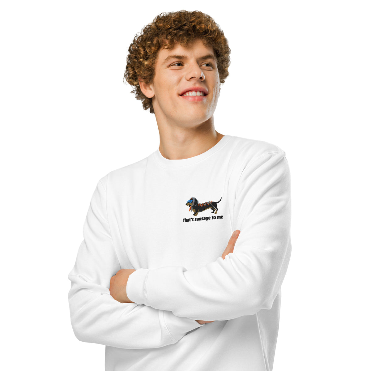 A smiling young man with curly brown hair wearing a white crewneck sweatshirt with a small dachshund graphic and the text 'that's sausage to me' on the left chest, posed with arms crossed against a solid white background.