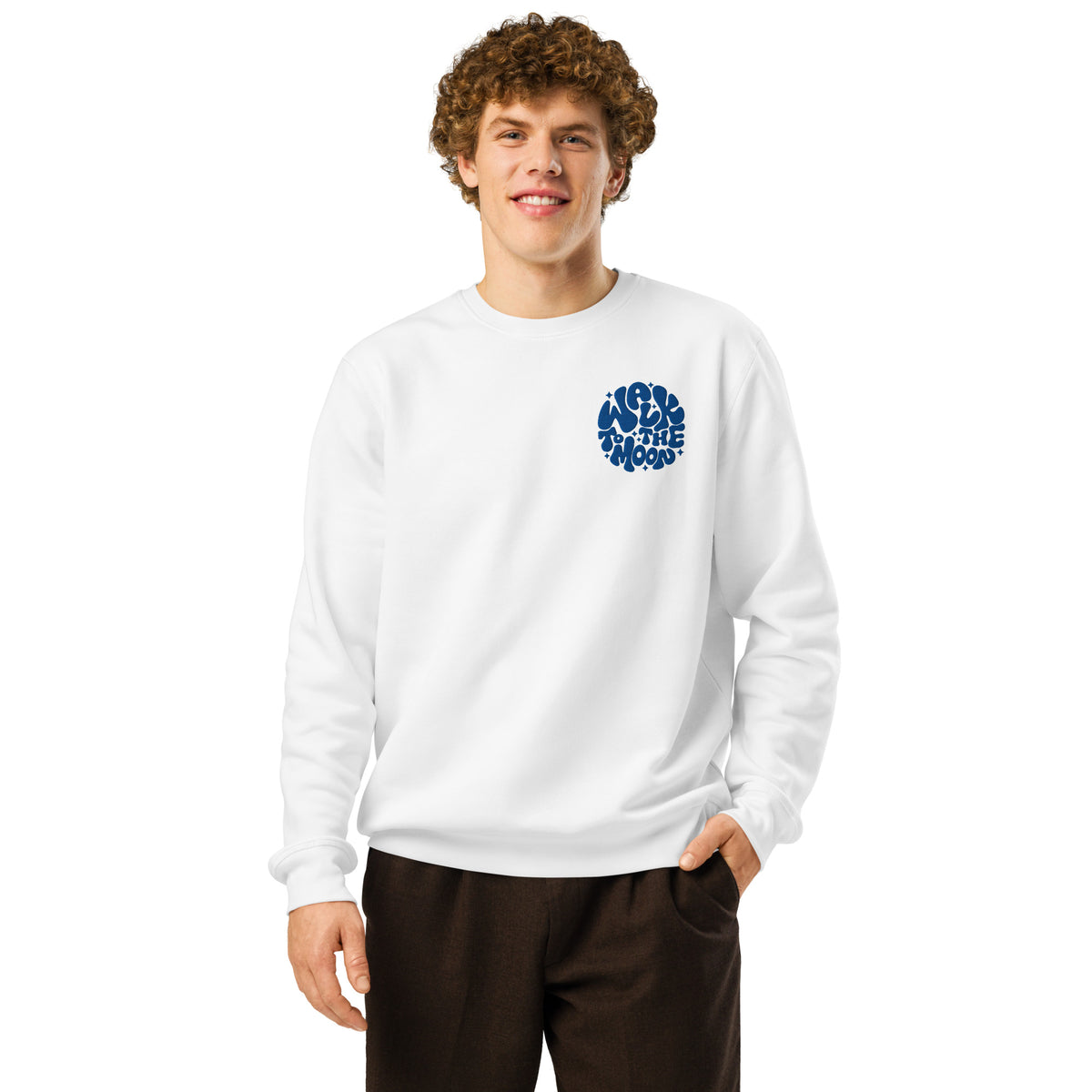 A man with curly brown hair wearing a white crewneck sweatshirt with a blue circular graphic on the left chest, paired with dark brown pants, against a solid white background.