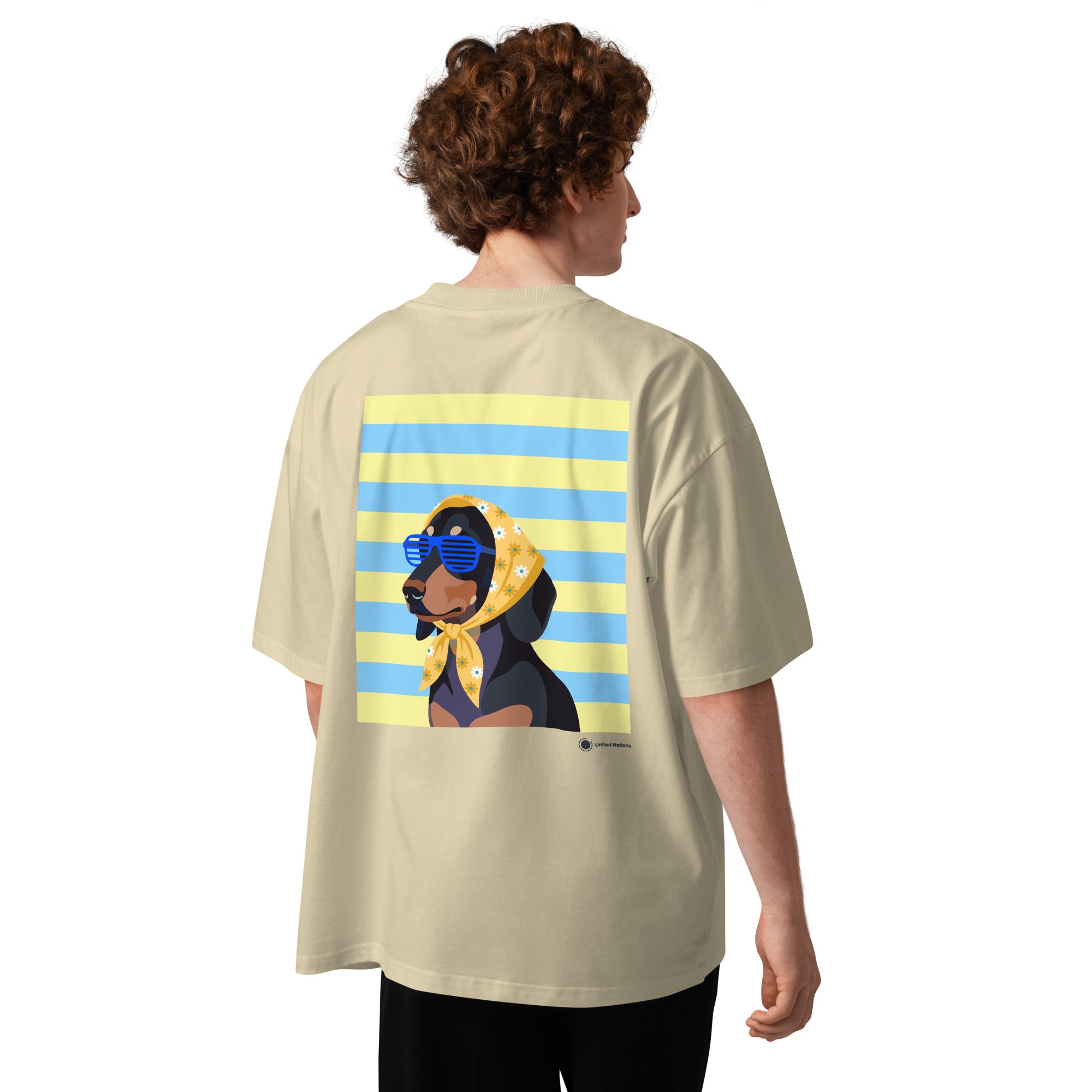 A person shown from the back wearing an oversized light beige t-shirt with a large graphic print of a dachshund wearing a yellow bandana and blue sunglasses, set against a background of yellow and blue horizontal stripes.