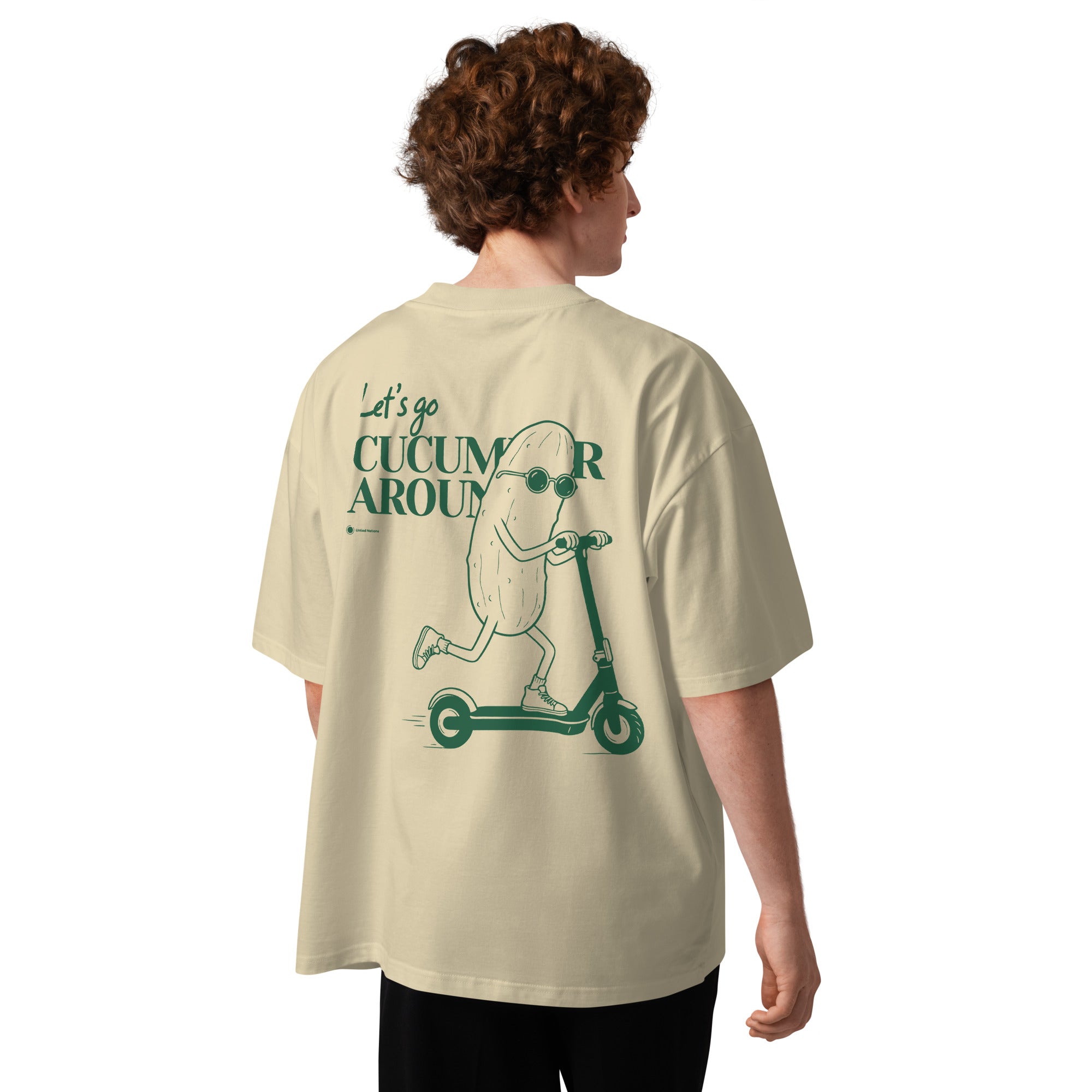 A person shown from the back wearing an oversized cream-colored t-shirt with a green graphic of a cucumber character riding a scooter and the text 'Let's go CUCUMBER AROUND'.