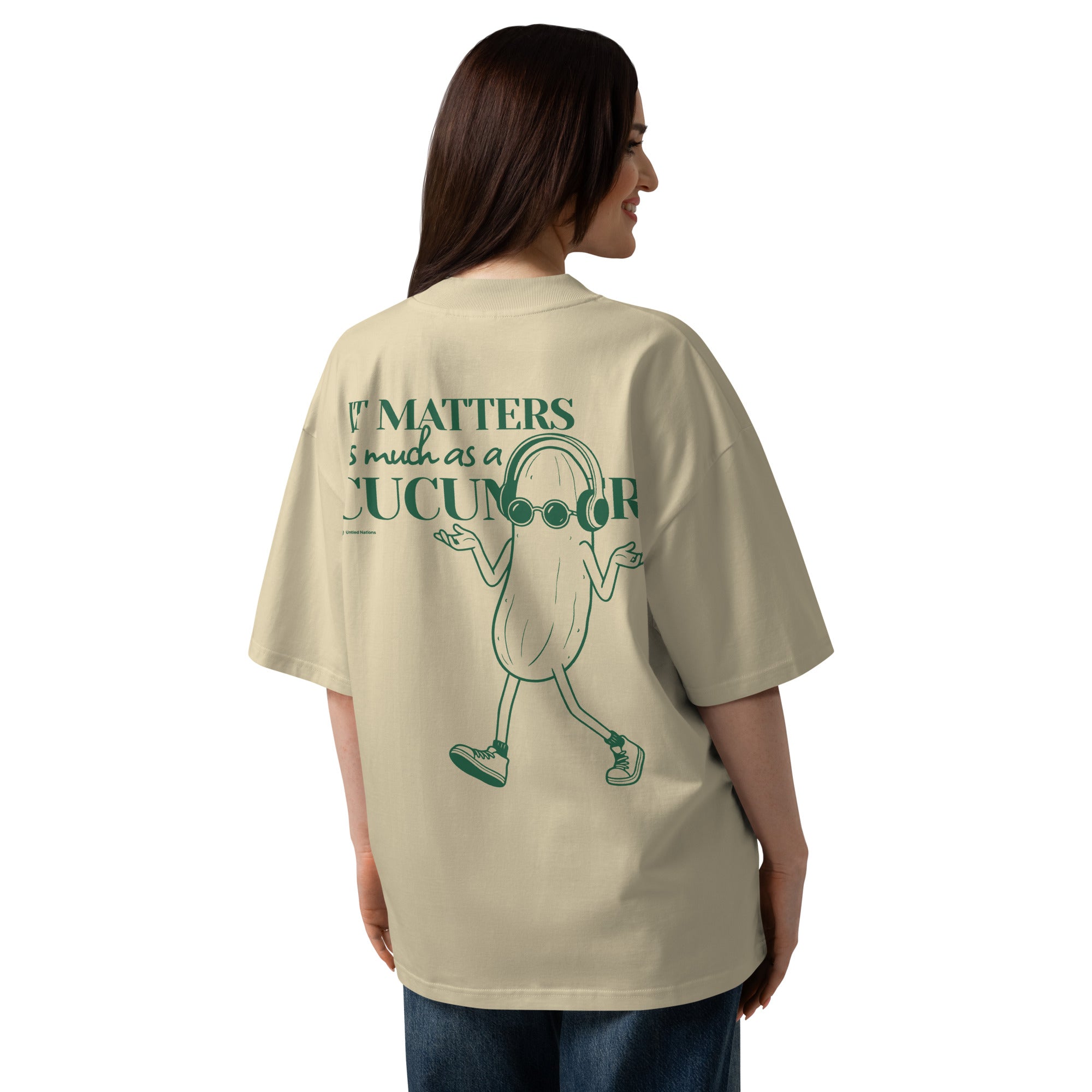 A woman shown from the back wearing an oversized beige graphic T-shirt with a green cartoon cucumber character and the text 'It matters as much as a cucumber'.