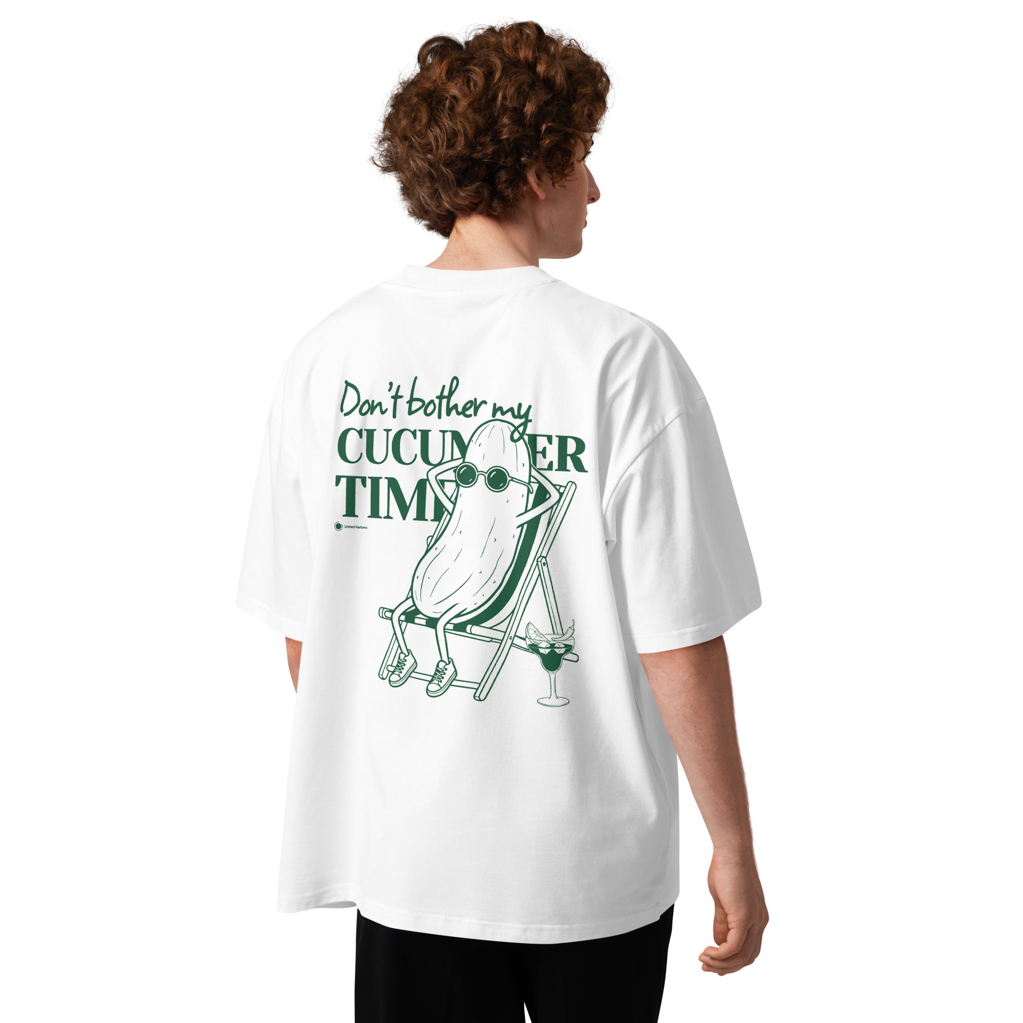 A person shown from the back wearing an oversized white T-shirt with a green graphic print that reads 'Don't bother my CUCUMBER TIME' and shows a cucumber relaxing on a beach chair. The model is photographed against a clean white background.
