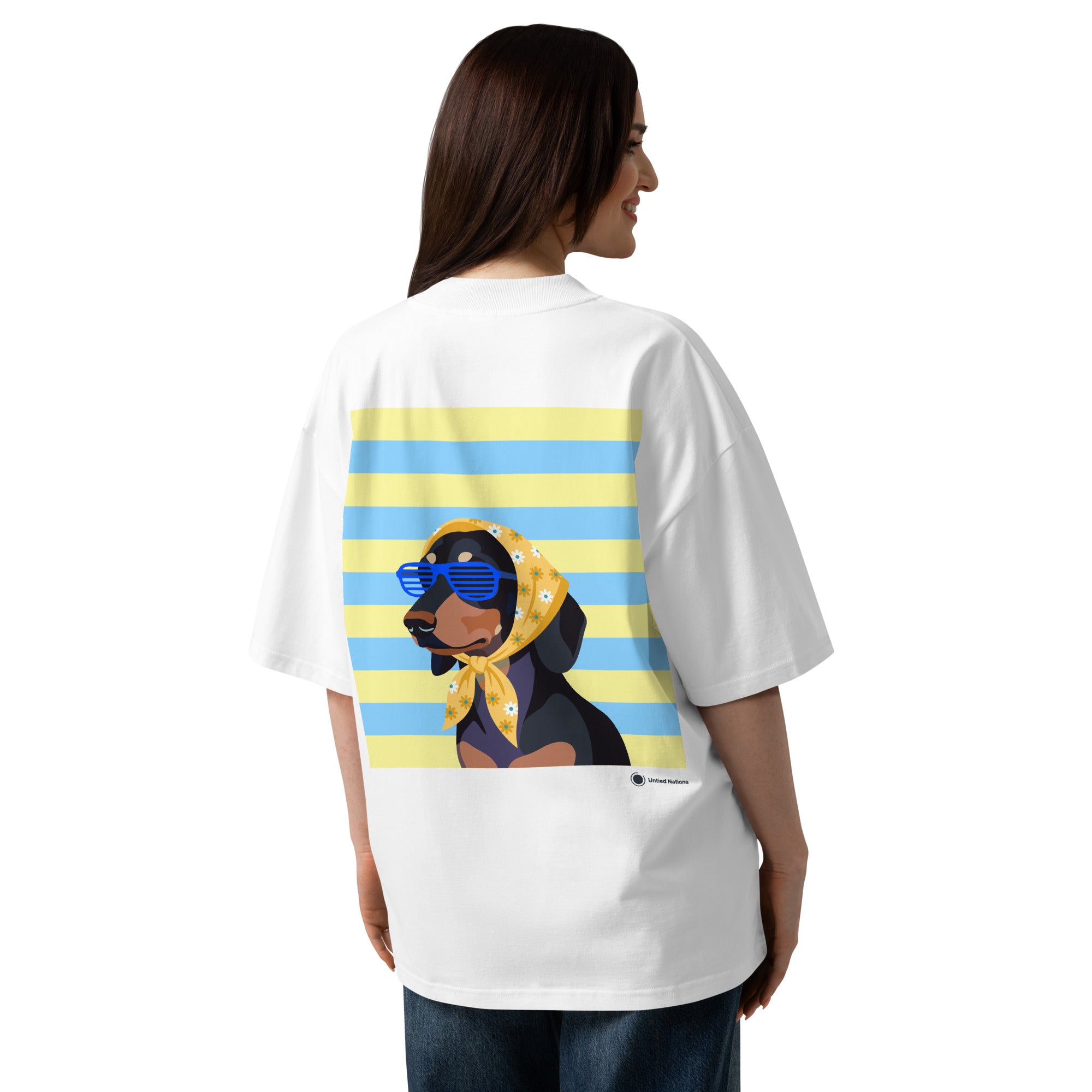 A woman shown from the back wearing an oversized white graphic T-shirt. The shirt features a large print of a dachshund wearing blue sunglasses and a yellow bandana, set against a background of light blue and yellow horizontal stripes.