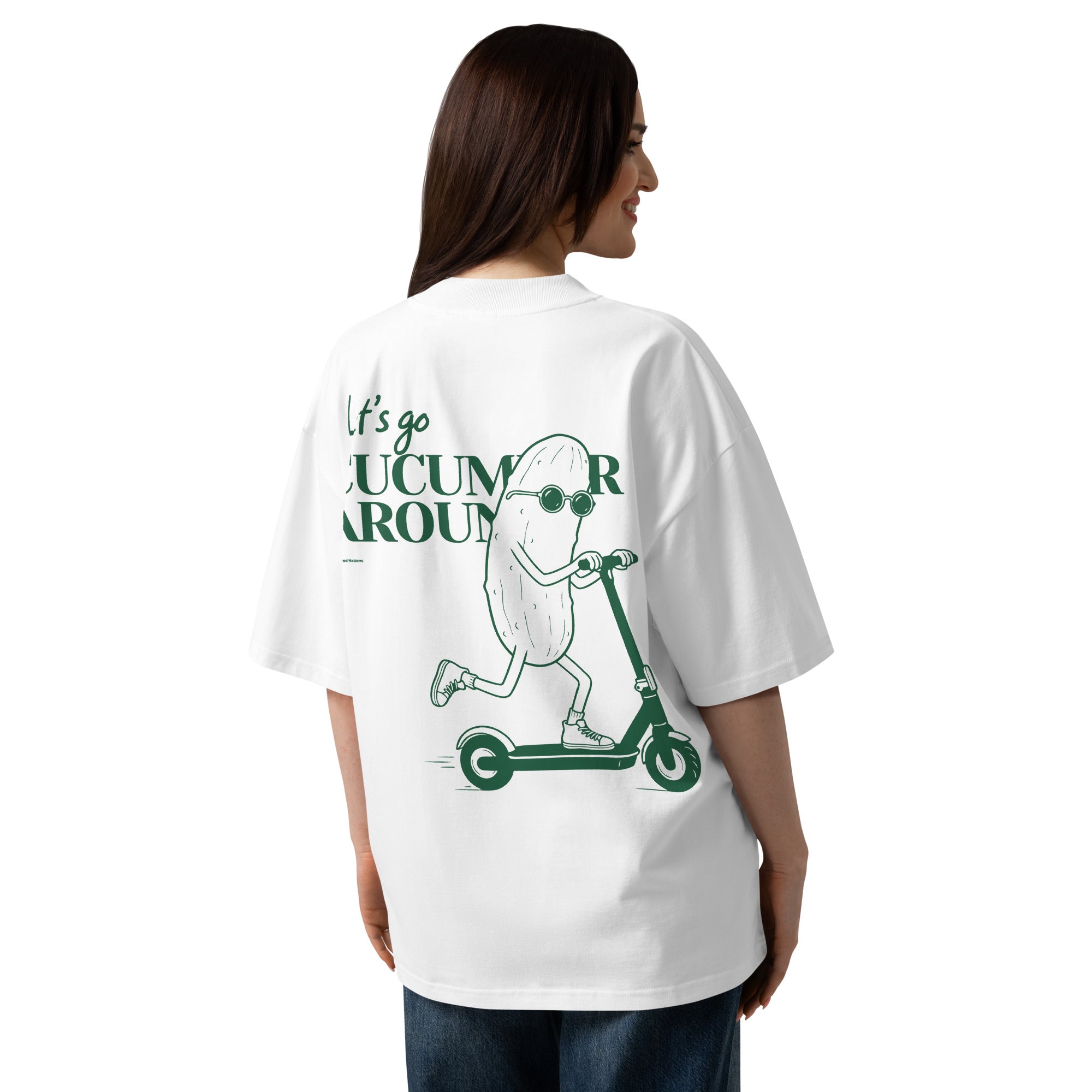 A woman shown from the back wearing an oversized white graphic T-shirt. The shirt features a green illustration of a cucumber character riding a scooter with the text 'It's go CUCUMBER AROUND'.