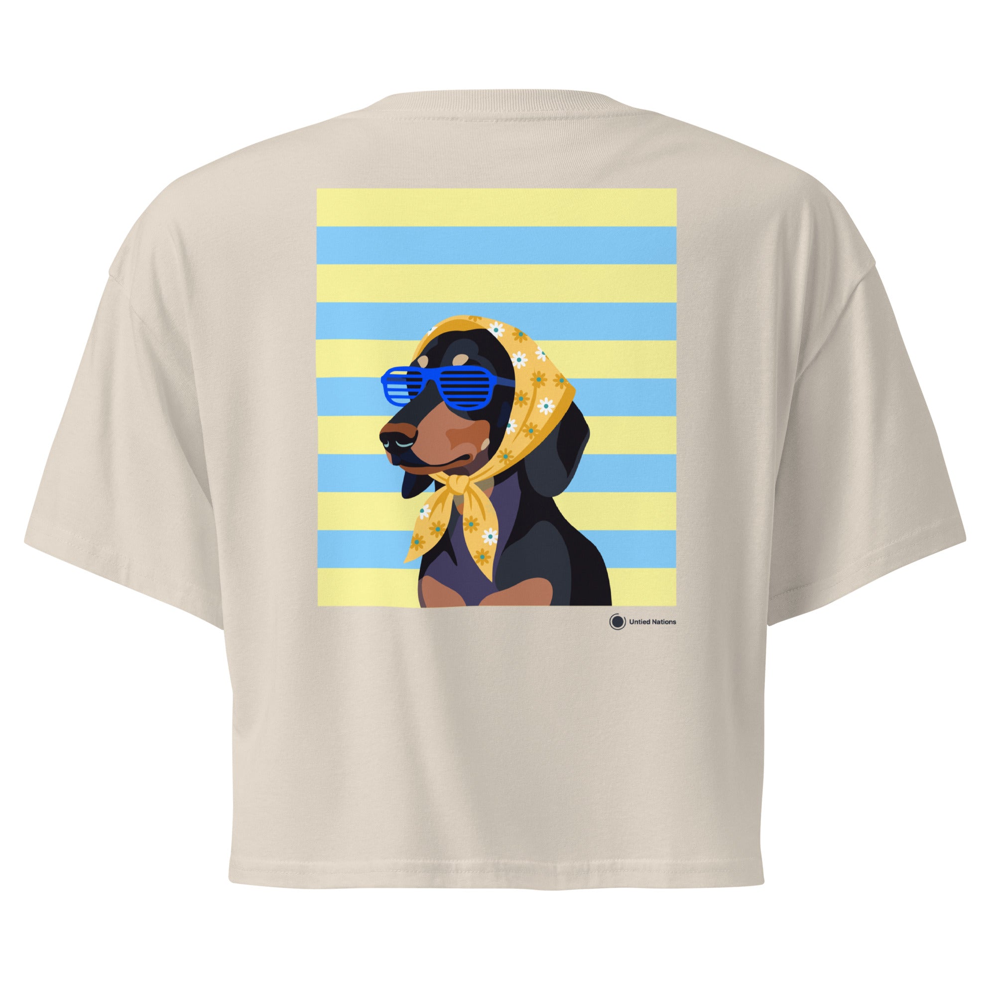 A beige short-sleeve crop top shown from the back, featuring a large graphic print of a black and tan dog wearing a yellow bandana and blue sunglasses, set against a background of yellow and light blue horizontal stripes.