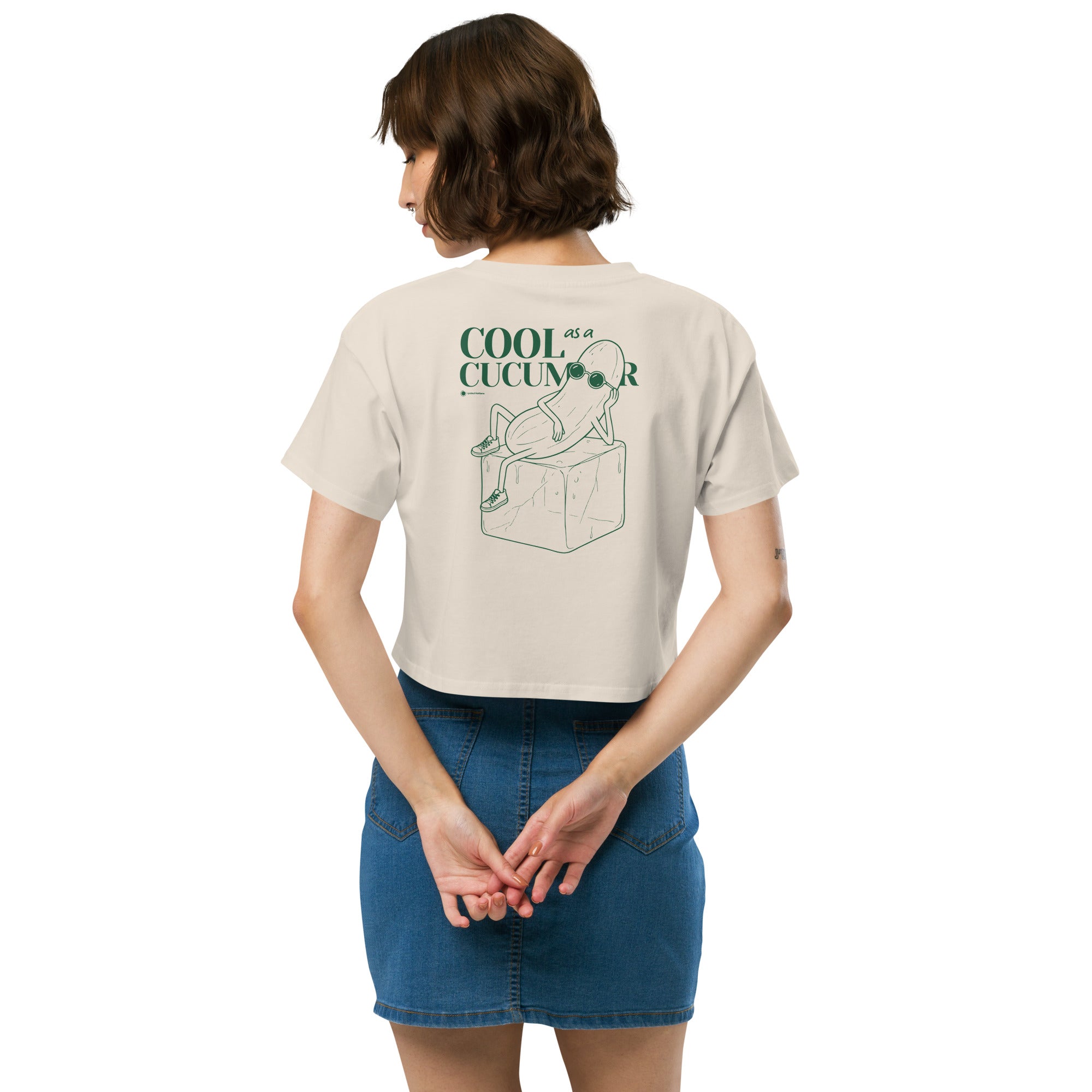 Person wearing a beige short-sleeve crop top shown from the back, featuring a green line art graphic of a cucumber character wearing sunglasses and sitting on an ice cube, with the text 'COOL as a CUCUMBER'.