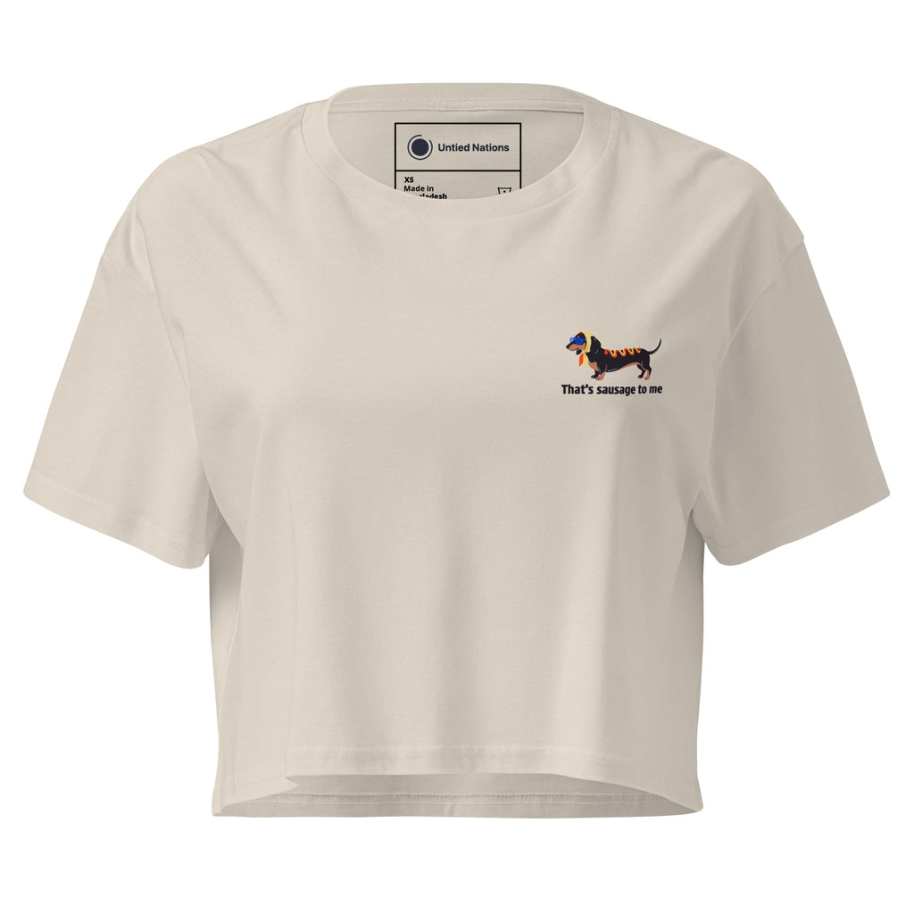 A cream-colored short-sleeve crop top t-shirt with a small graphic of a dachshund on the left chest and the text 'that's sausage to me' underneath, displayed on a clean white background.
