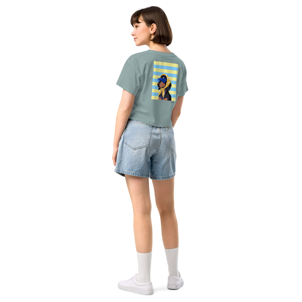 A woman shown from the back wearing a sage green t-shirt with a graphic print on the back, paired with light blue denim shorts, white socks, and white sneakers, set against a plain white background.