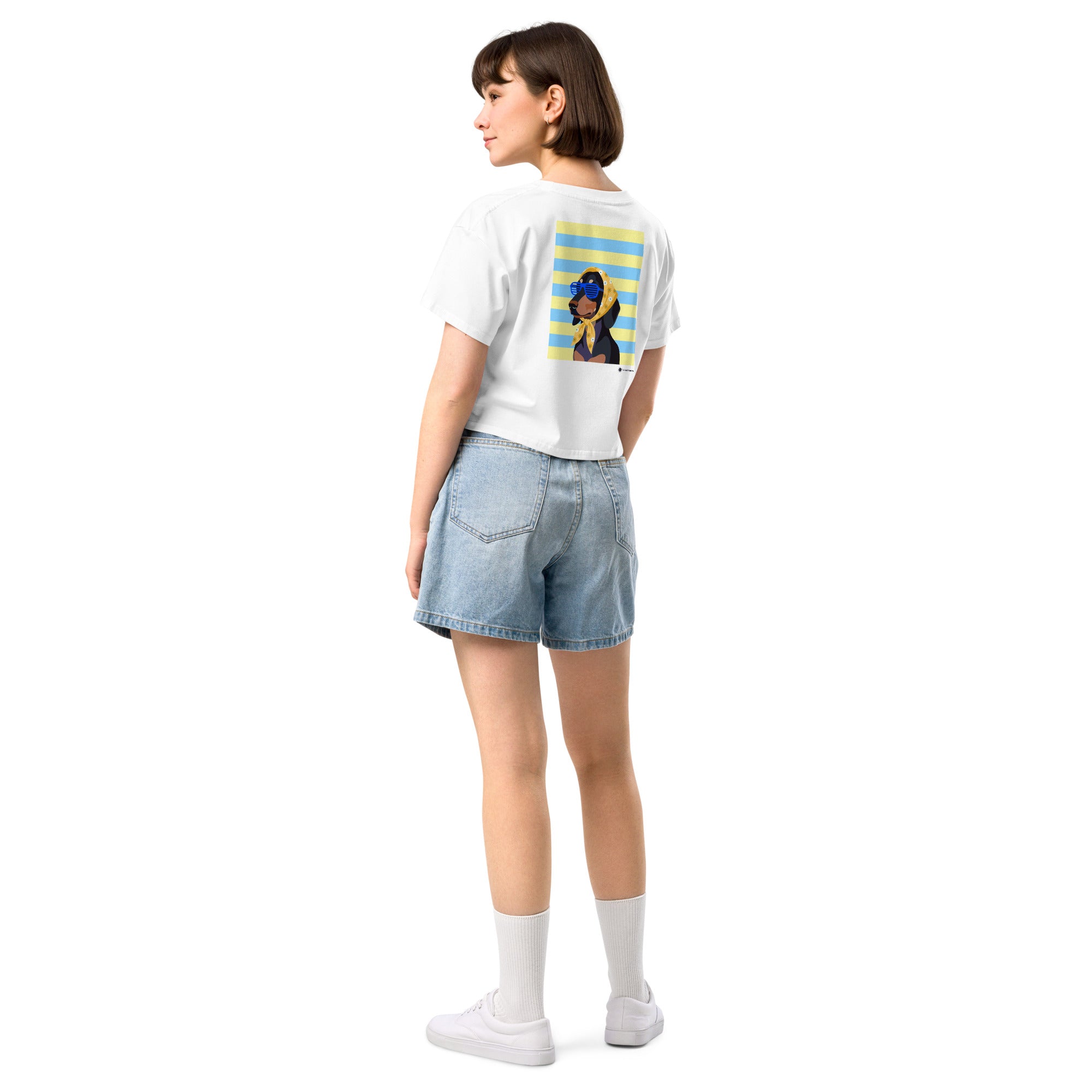 A woman shown from the back wearing a white graphic t-shirt with a colorful print on the back, paired with light blue denim shorts, white socks, and white sneakers, set against a plain white background.