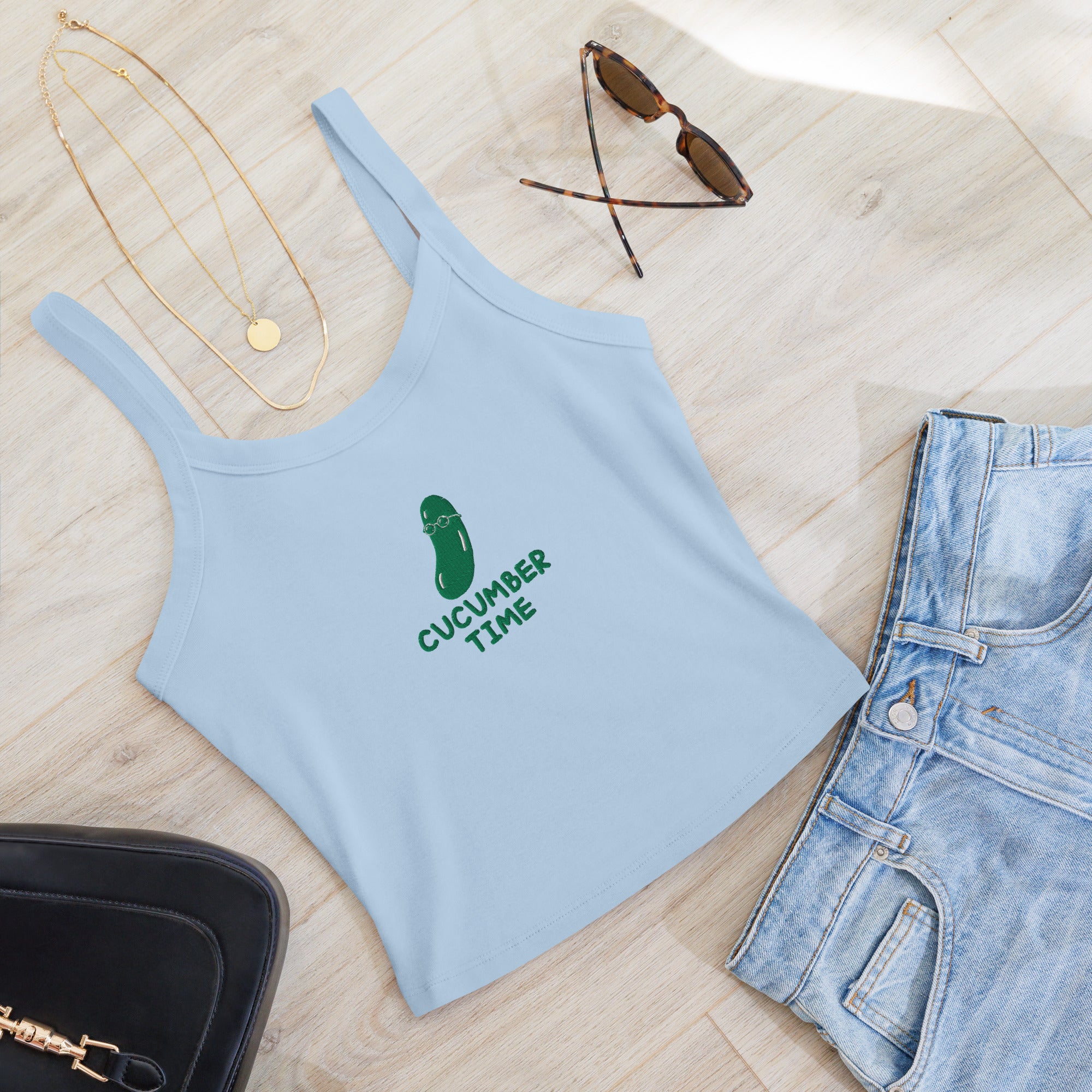 Light blue tank top with a logo, sunglasses, necklace, and denim shorts on a wooden surface.