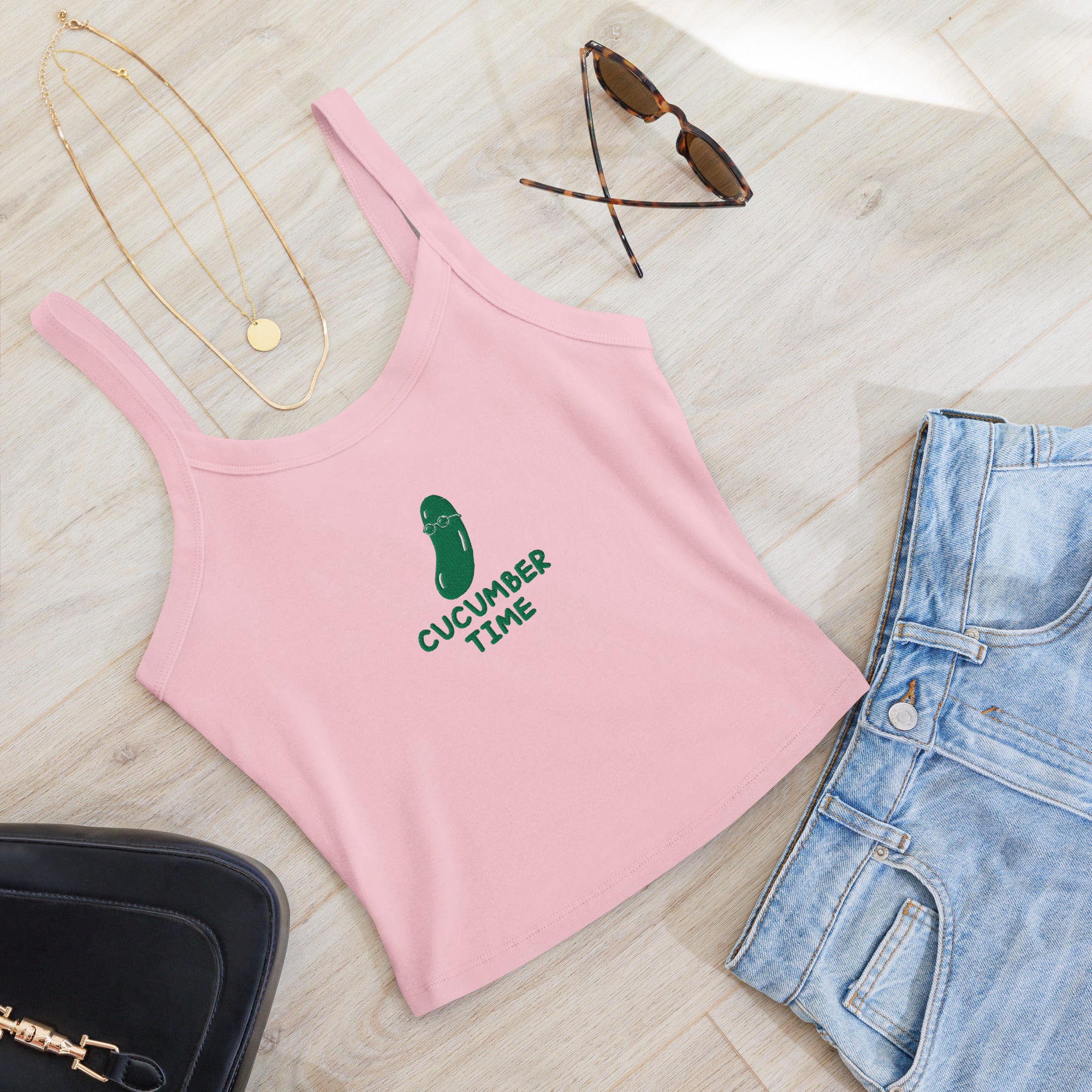 A pink ribbed tank top with a green cucumber graphic and the text 'CUCUMBER TIME' laid flat on a light wood floor, styled with blue jeans, sunglasses, gold necklaces, and a black handbag.