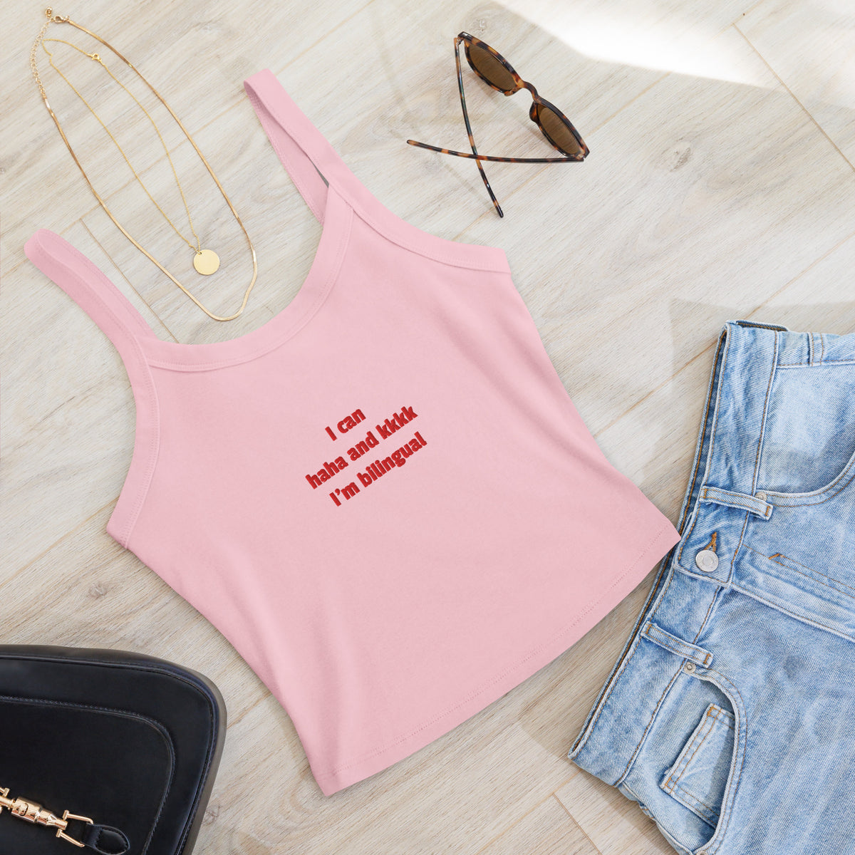 A pink ribbed tank top with red text reading 'I can haha and kkkk I'm bilingual' is styled in a flat lay on a light wood floor with light-wash jeans, gold necklaces, tortoiseshell sunglasses, and a black handbag.