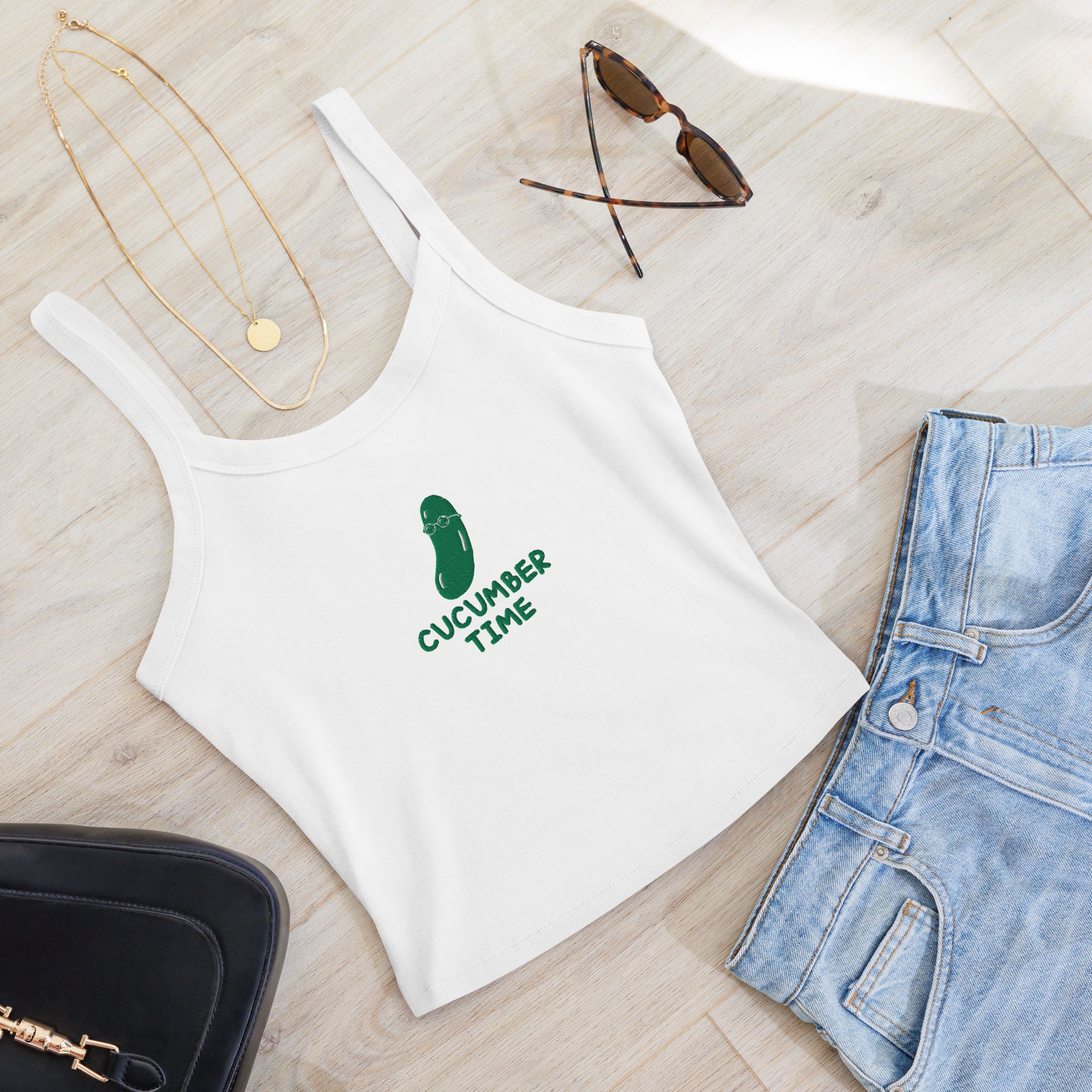 A white ribbed tank top with a green cucumber graphic and the text 'CUCUMBER TIME' laid flat on a light wood floor, styled with light blue jeans, sunglasses, gold necklaces, and a black handbag.