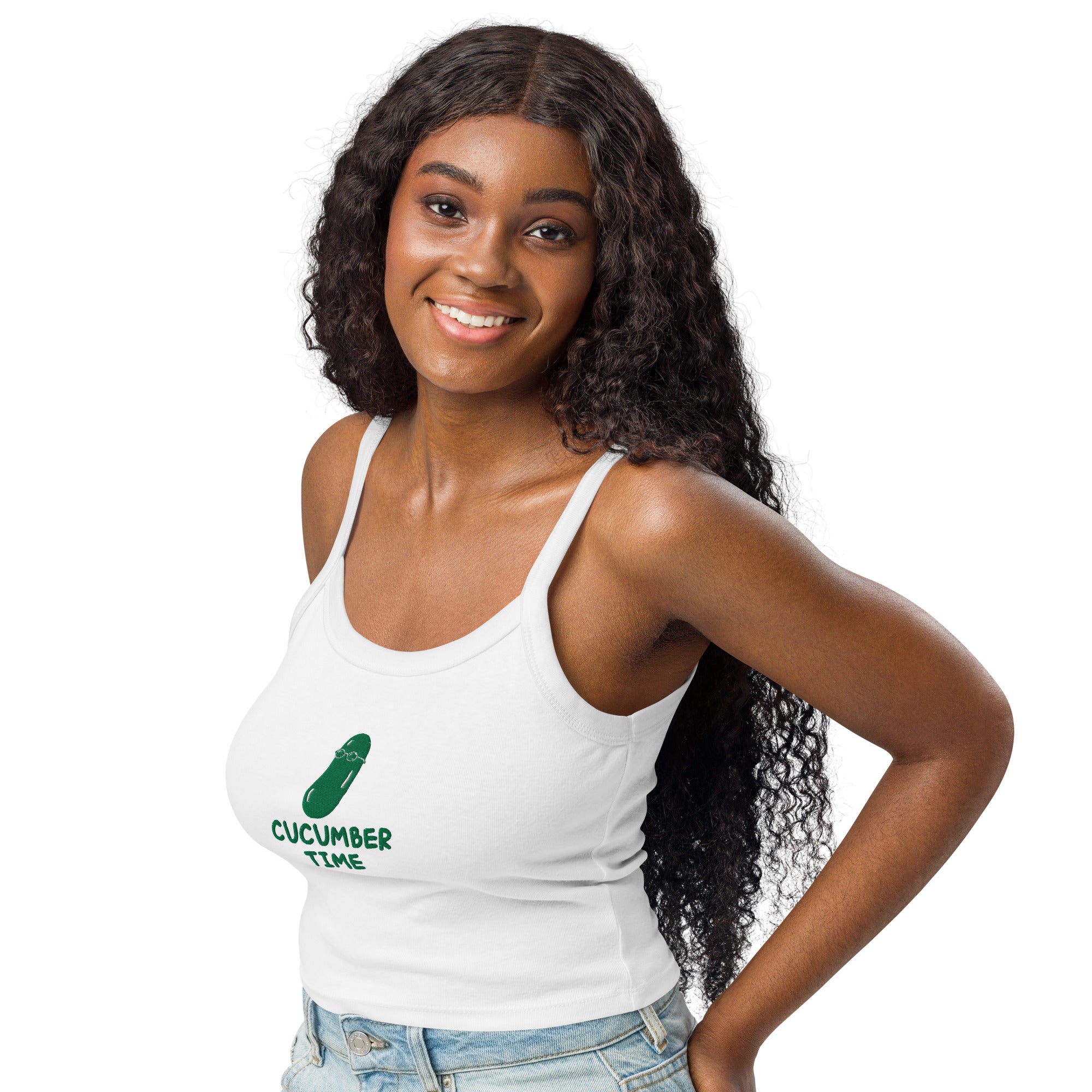 A smiling woman with long, curly dark hair models a white ribbed tank top featuring a green cucumber graphic and the text 'CUCUMBER TIME' on the front, paired with light-wash denim jeans against a plain white background.