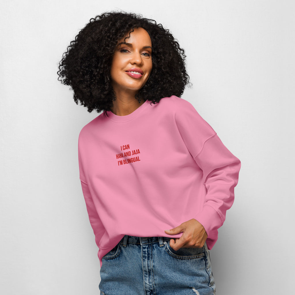 A woman wearing a pink crewneck sweatshirt with the text 'I can haha and jaja I'm bilingual' printed on the chest, paired with blue jeans, against a plain light gray background.
